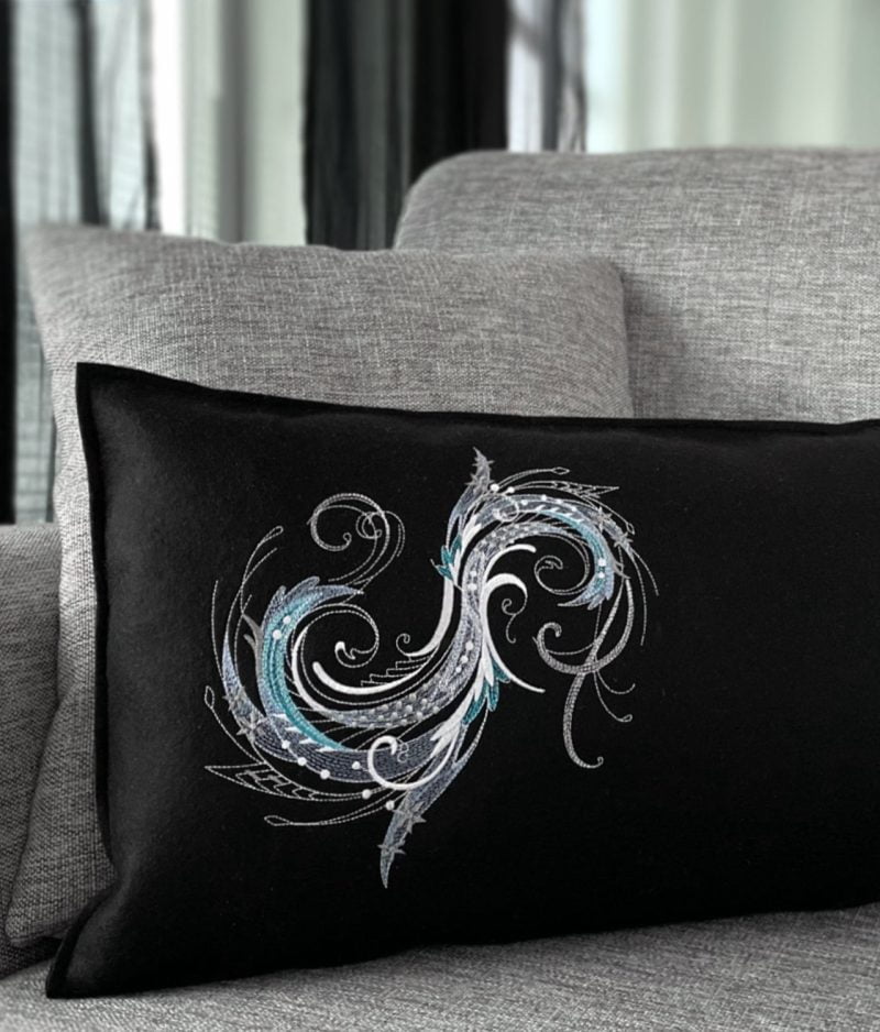 Harmaalla sohvalla mustapohjainen villahuovasta valmistettu tyyny, jossa konekirjottu valkoisen ja turkoosin sävyinen pyörre-kuvio.