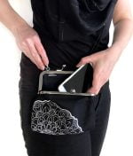 Musta kukkarolaukku, jossa valkoinen mandala-kuvio, naisen olalla. Nainen laittaa puhelimen laukkuun.
