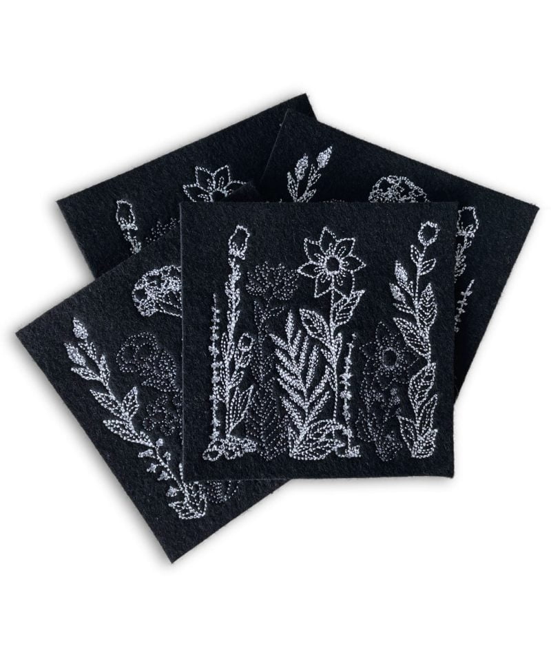 Neljä mustaa huovasta käsityönä valmistettua lasinalusta, joissa konekirjottu valko-harmaa kukkaketo-kuviointi.