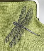 Tummanharmaalla metallilangalla konekirjottu lentävä sudenkorento laukun etupuolella vihreällä kankaalla.
