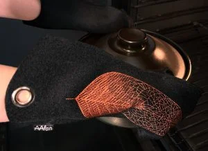 Mustasta villahuovasta käsityönä valmistetut patakintaat, joissa kuparinvärinen brodeerattu lehti-kuvio, naisen käsissä laittaessa mustaa uunipataa uuniin.