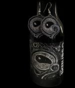 Ikiiurso-korvakorut roikkumassa mustapohjaisen Hiisin Ikiiurso-pullon kyljessä etiketin yläpuolella.