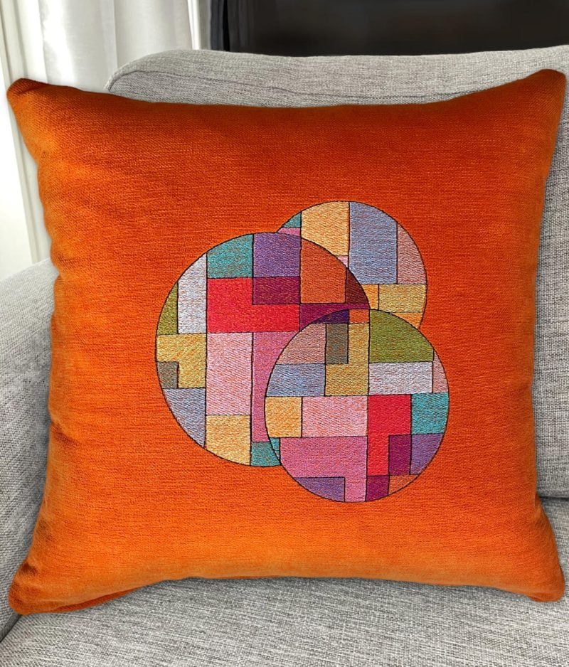 Harmaalla sohvalla oranssi pehmeäpintainen tyyny, jossa brodeerattuna värikkäistä suorakulmioista muodostuvat kolme ympyrää yhteensommiteltuna.