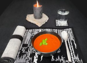Tummataustainen kuva kauttauksesta, jossa musta kulho punertavaa tomaattikeittoa aseteltuna mustapohjaiselle villahuovasta valmistetulle tabletille, jossa valkoinen konekirjottu koivumaisema.