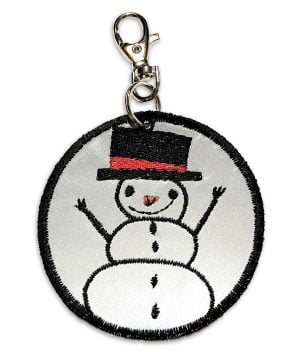 Heijastinkankaasta käsityönä valmistettu pyöreä riipus, johon konekirjottu mustalla viskoosilangalla silinteripäinen, hymyilevä lumiukko.