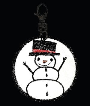 Kuva heijastinkankaasta käsityönä valmistetusta riipuksesta pimeällä, jossa mustalla viskoosilangalla konekirjottuna silinteripäinen, hymyilevä lumiukko.