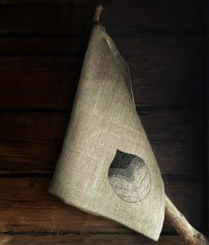 A cozy picture of a linen towel with a black embroidered leaf hanging in the hook on a log wall.