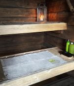 Linen Leaf-cloth with green embroidered leaf in the corner on a lower bench in a cozy log sauna. Green shampoo bottles and a black shower brush beside it. On the upper bench a lantern with a lit candle inside creating atmospheric feeling.