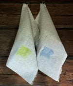 Handcrafted two linen towels hanging on a log wall. In the other towel machine embroidered green leaf in the corne, and the other, a gray emroidered leaf.