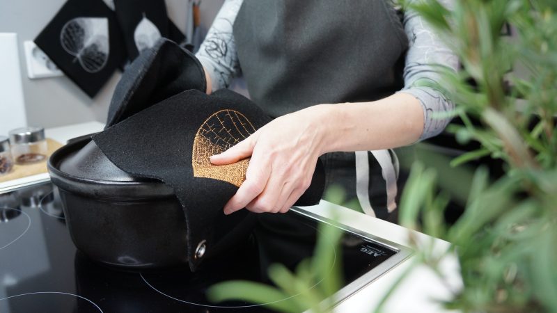 Etualalla kuvassa sumea rosmariinin oksa. Kuva tarkentuu liedelle laskettuun mustaan keraamiseen uunipataan, jonka reunasta nainen pitelee kiinni käden suojana musta villahuovasta valmistettu patalappu pronssinvärisellä Lehti-kuviolla. Toisessa kädessä näkyy taaempana musta villahuopainen patakinnas. Taustalla harmaalla seinällä roikkuvat valkoisella konekirjotuin kuvioin Lehti-patalappu ja -kinnas.