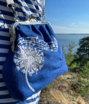 Sinivalkoraitaiseen vaatteeseen pukeutuneella naisella käytössä sininen verhoilun ylijäämävillakankaasta valmistettu kehyskukkarolaukku. Laukussa valkoisella konekirjottu voikukan pölyttynyt kukinto, josta tuulen mukana lentää nuotteja, muodostaen kaarevan nuottiviivaston. Kuvassa taustalla sininen taivas ja merimaisema.