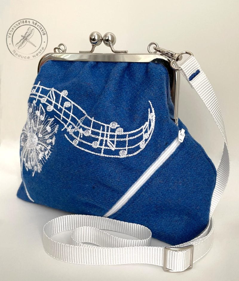 Blue purse handcrafted from remnant woolfabric pictured from right front view. In the picture a white closed zip pocket declining to the right. On the middle part of the purse embroidered notes on the staff.