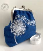 Blue Melody Purse handcrafted form remnant woolfabric pictured from the left front view. On the front clear image of the fluffy dandelion machine embroidered with white and light gray rayon thread. In between of the fluffy seeds notes and a clef.