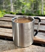 Metallinen, kahvilla täytetty, Lyhty-retkimuki puisella pöydällä. Taustalla näkyy kuusimetsää.