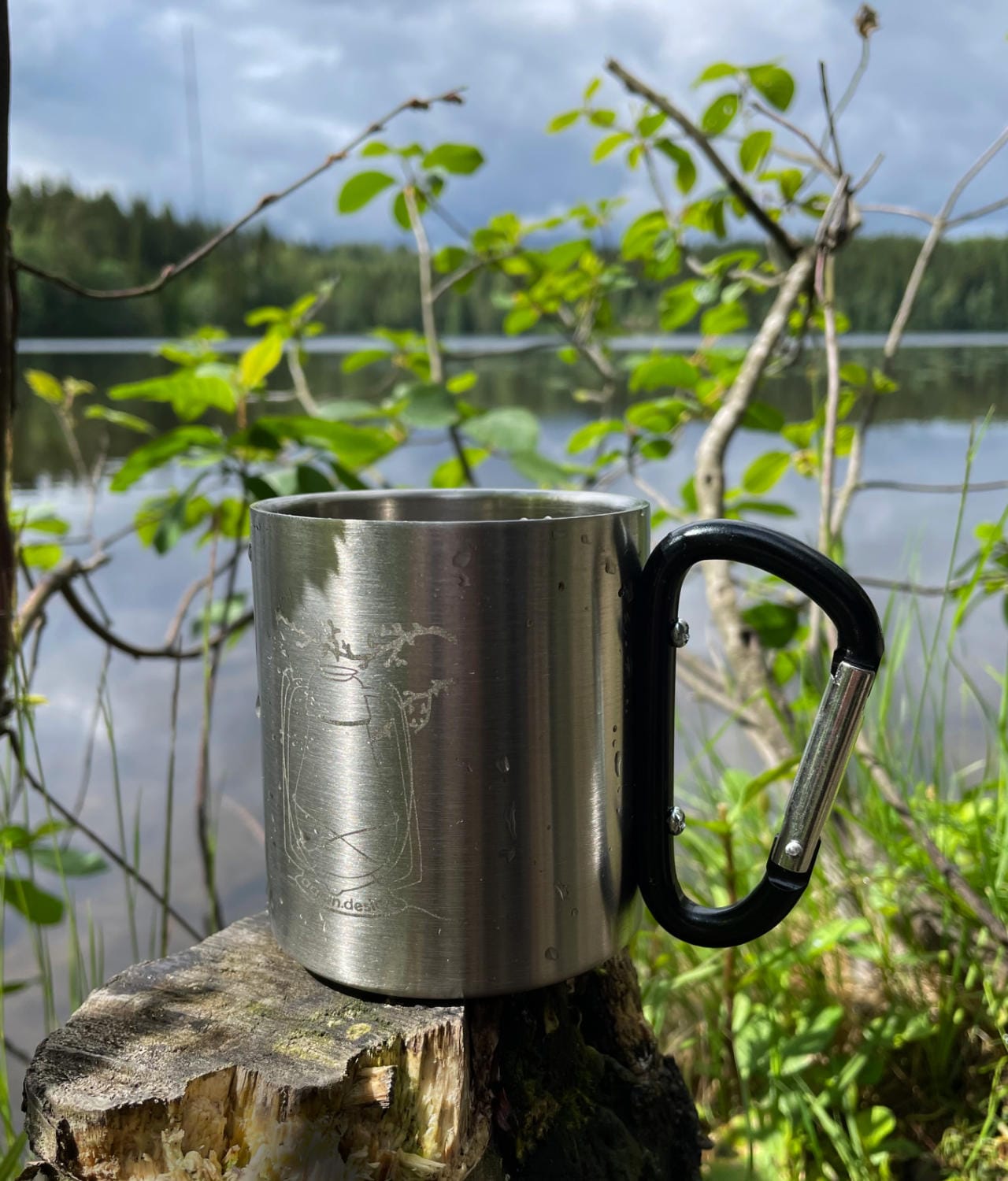 Tuotekuvat Lyhty-muki kannon päällä. Taustalla järvimaisema ja vehreitä oksia.