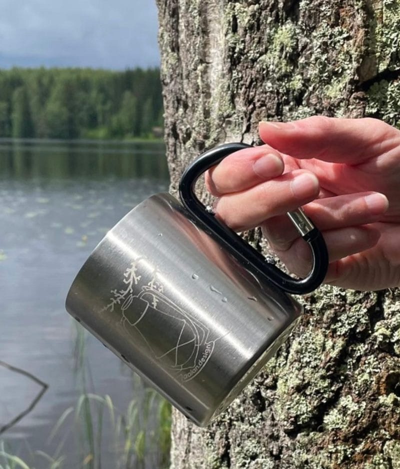 Metallinen Lyhty-muki rennosti naisen sormissa roikkumassa karabiinikahvasta. Taustalla osittainen puunrunko ja järvimaisema.