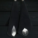 Kaksi mustaa vohvelipintaista kasvopyyhettä roikkkumassa mustalla puupaneliseinällä valkoisista koukuista. Pyyhkeissä konekirjotut valkoiset Lehti-kuviot alakulmassa.