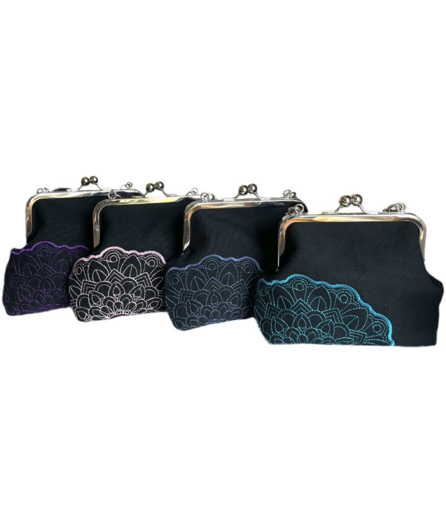 Neljä mustapohjaista käsityönä valmistettua kukkarolaukkua, joissa viskoosilangalla konekirjottu pitsimäinen mandalakuvio eri väreissä.