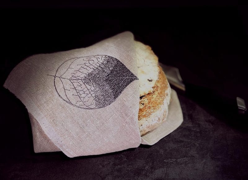 Mustataustainen kuva, jossa vastapaistettu leipä kiedottu Lehti-kuvioituun pellavaliinaan. Mustalla kirjottu lehtikuvio jää kauniisti näkyviin.