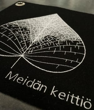 Musta villahuovasta valmistettu pannulappu kuvattu viistosti edestä, jolloin etualalla konekirjottu esimerkki nimikointitekstistä (”Meidän keittiö”).