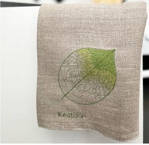 Vihreällä konekirjottu lehtikuvio ja nimikointiteksti ”Keittiöön” pellavaisessa liinassa. Taustalla hahmottuu keittiön välineitä.