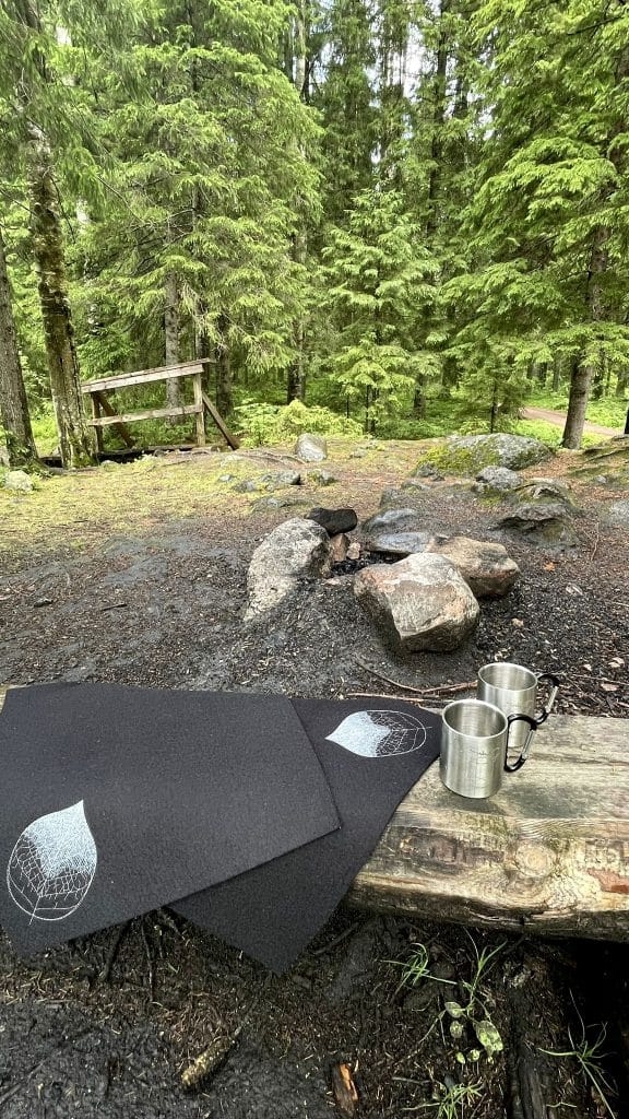 Kuva metsäiseltä retkeltä sammutetun nuotion ääreltä. Etualalla hirsipenkillä mustat villahuopaiset Lehti-istuinaluset ja rosteriset Lyhty-retkimukit.