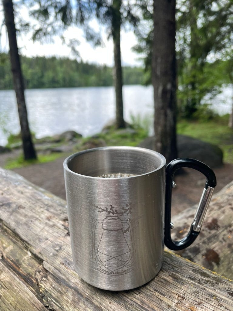 Rosterinen Lyhty-muki, jossa kahvia, asetettu hirsiparrun päälle. Taustalla metsäinen järvimaisema.
