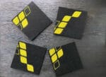 Pöydällä 4 lasinalusta, joista muodostuu oikein päin yhteen aseteltuna Hiisin keltainen logo.