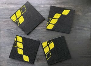 Pöydällä 4 lasinalusta, joista muodostuu oikein päin yhteen aseteltuna Hiisin keltainen logo.