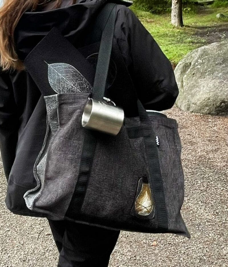 Musta saunakassi, jossa konekirjottu myrskylyhty naisen olalla retkikäytössä metsässä.