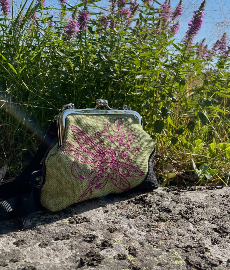 Vihreätaustainen verhoilun ylijäämäkankaista valmistettu laukku konekirjotulla pinkillä sudenkorentokuviolla kalliolla, jonka jäkälät ja taustalla näkyvät kesäiset pinkit kukat ovat innoittaneet laukun väriyhdistelmään.